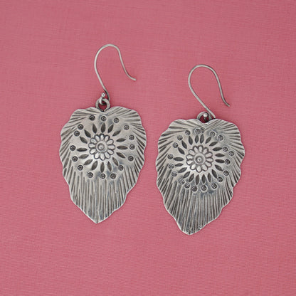 Silver leaf-shaped earrings with intricate designs on a pink background